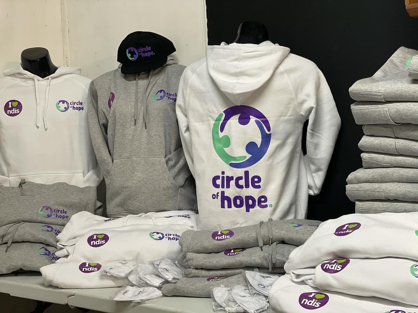 White and grey Circle of Hope hoodies and a black cap with the provider logo, folded in a stack