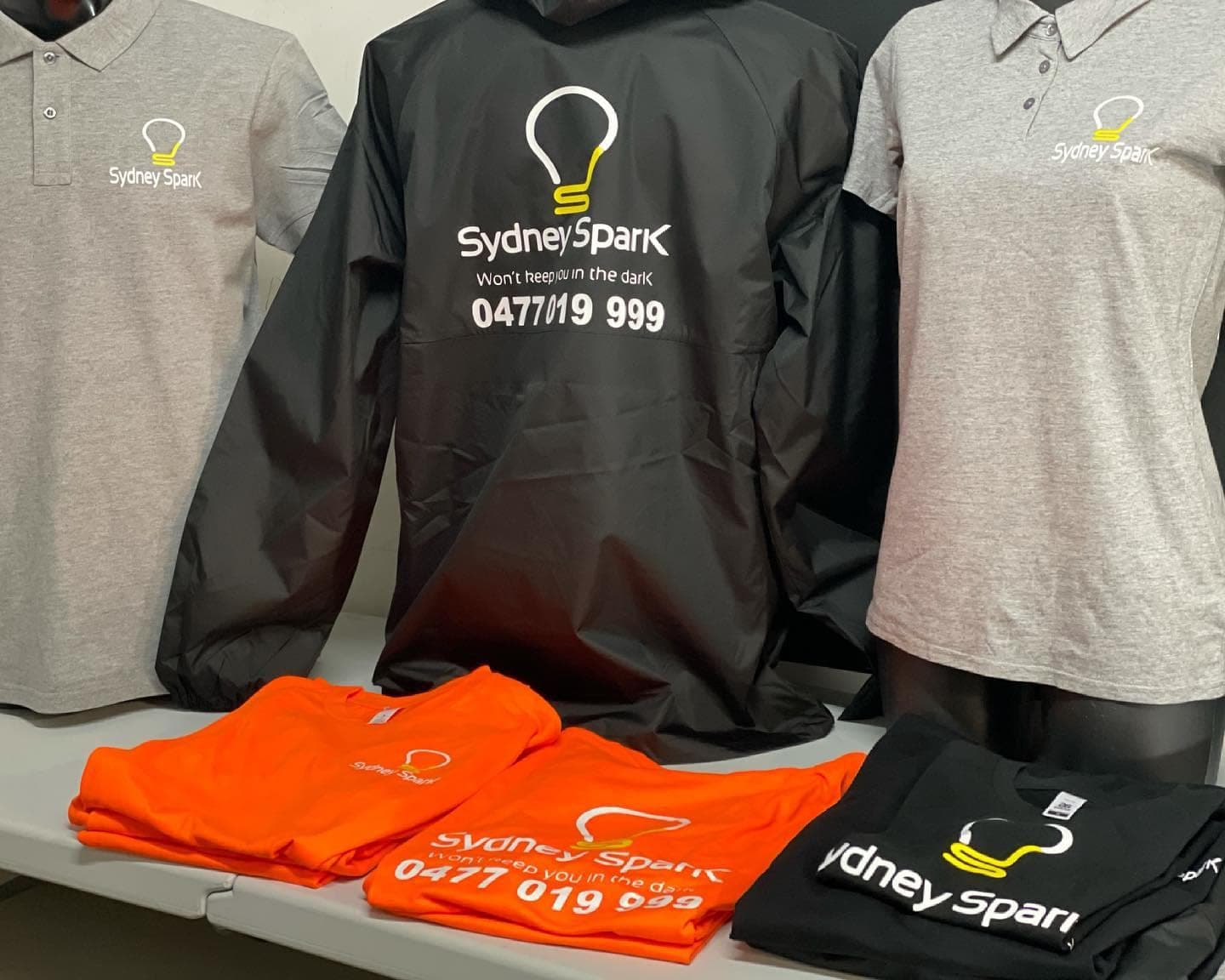 Grey Sydney Spark Services polos, a black jacket with a large back print and orange tees laid out as a uniform kit